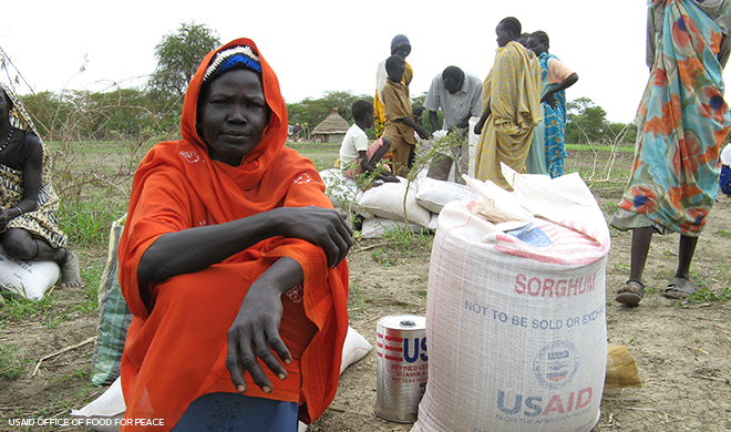 woman sits next to USAID bag of supplies while other people stand and walk nearby